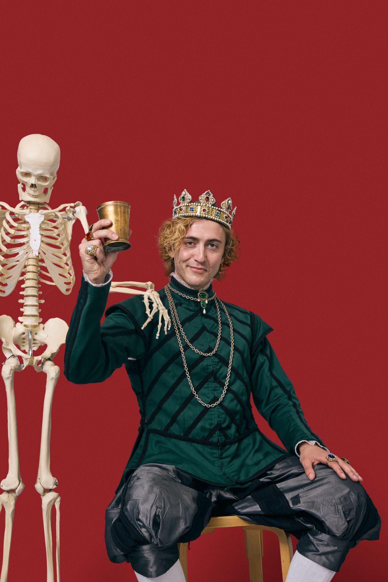 Mand i grønt renæssancekostume med guldkrone holder et gyldent bæger ved siden af et skelet – teatralsk foto til Hamlet-aktiviteter på Kronborg
