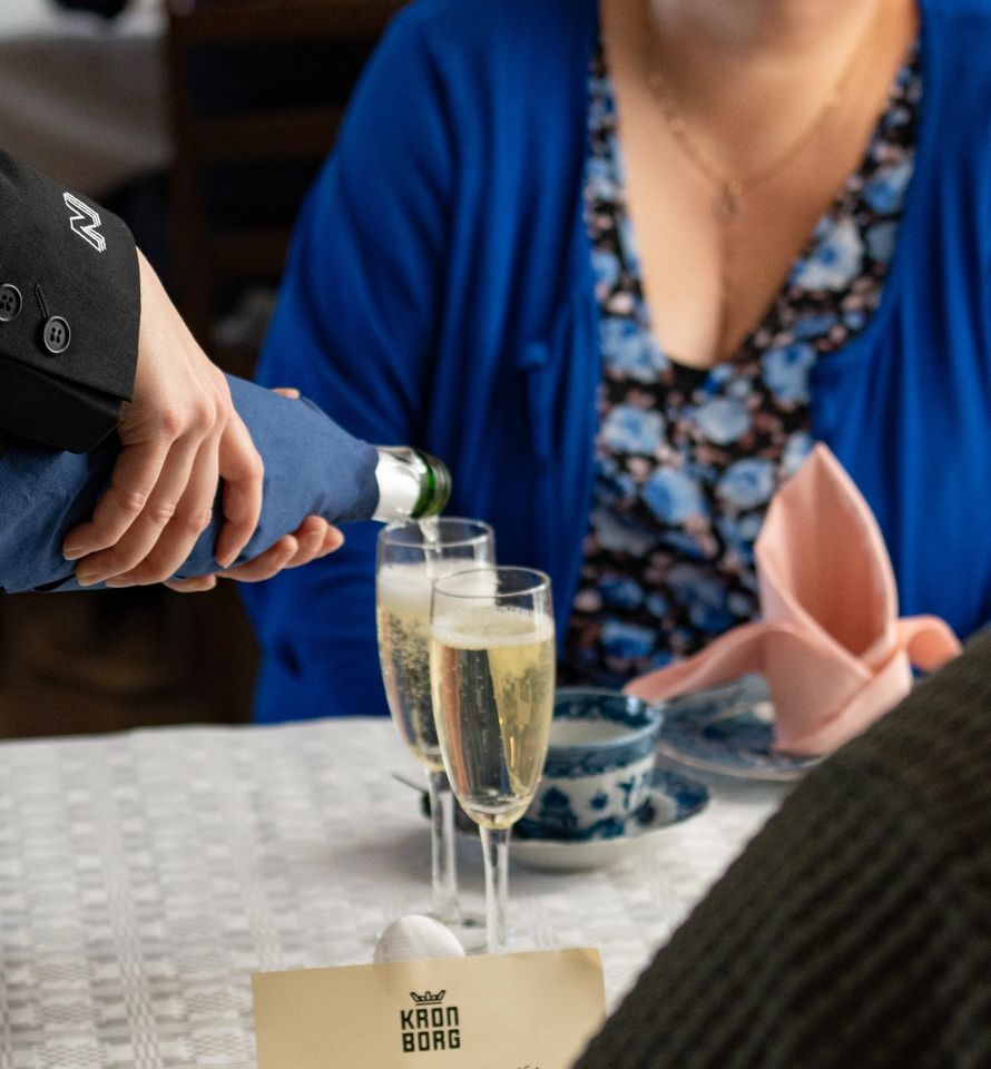 Champagne hældes i glas ved afternoon tea-bord med Kronborgs menukortet synligt – nærbillede der viser den elegante velkomst til arrangementet