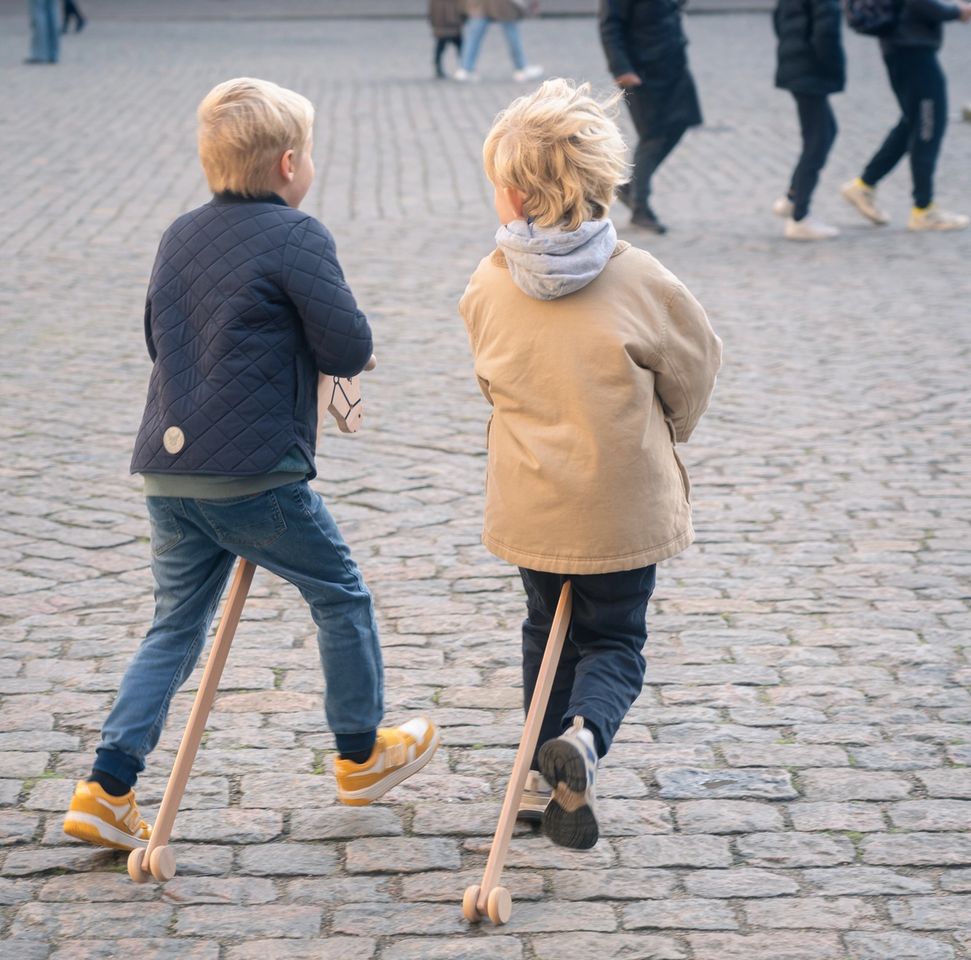 To drenge løber på kæpheste hen over Kronborgs brolagte gårdsplads med besøgende i baggrunden – historiske legeaktiviteter for børn på slottet